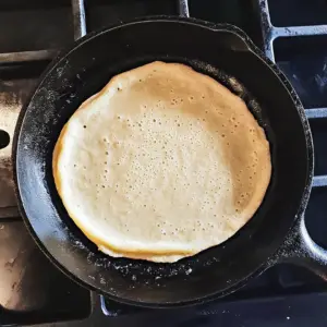 cast iron skillet with a handle on a stovetop with a scoop of millet pancake batter sizzling in the center